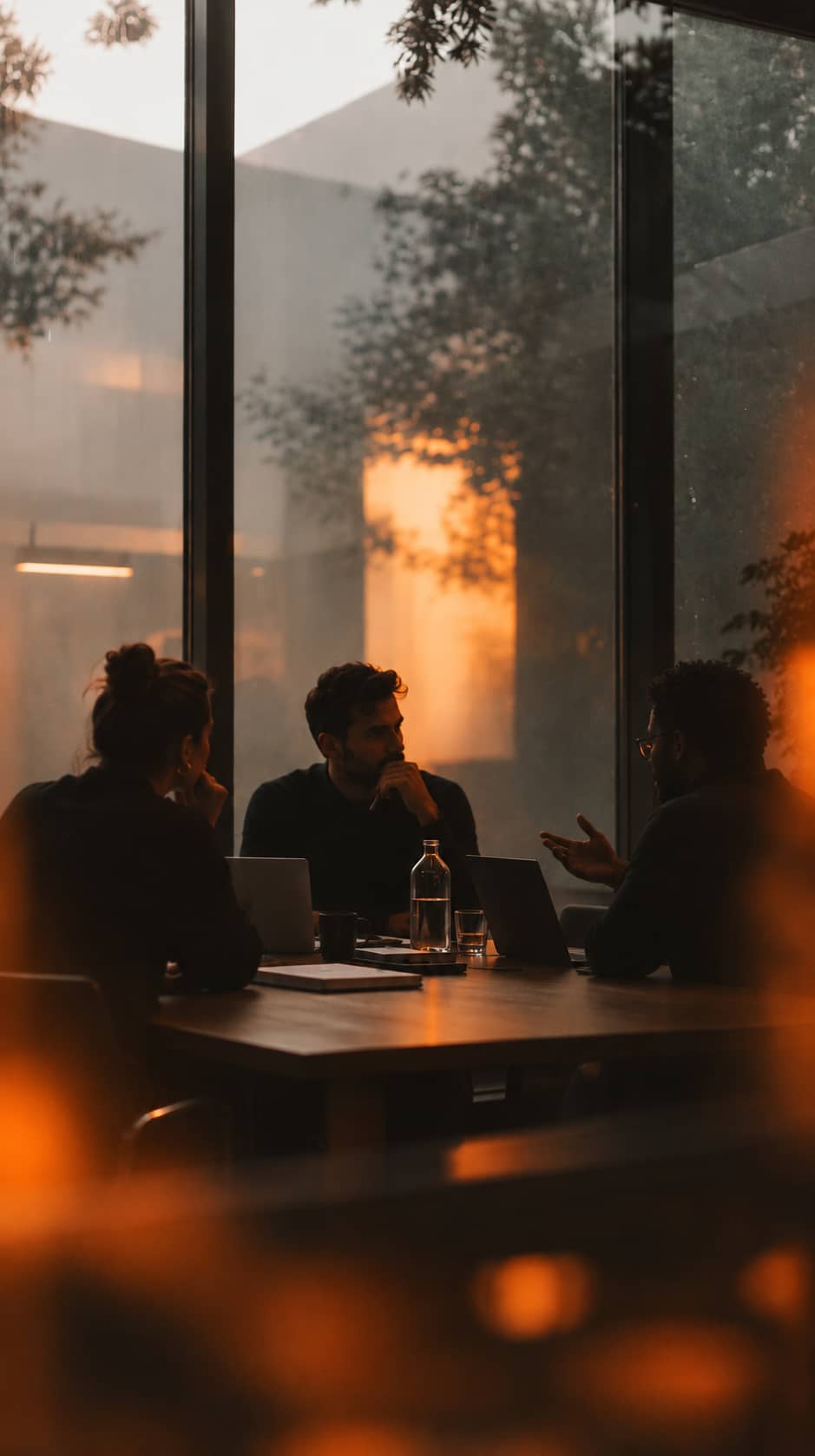 Three colleagues meeting around a table in a warmly lit office at dusk